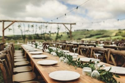 Outdoor wedding withhired wooden tables, wooden cross back chairs, greenery garlands cutlery and plates