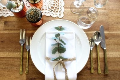 wedding place setting with hire glasses, brass handeled cutlery and crockery on a rustic wooden table with linen napkin and gum decoration